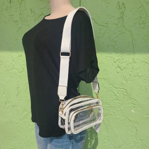 May include: A clear crossbody bag with white trim and a white adjustable strap. The bag has three zippered compartments with gold-colored zippers. The bag is worn over the shoulder.
