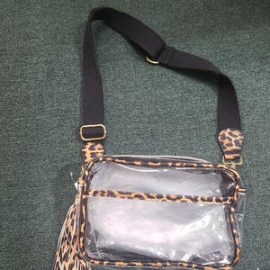 Puede incluir: Bolso cruzado transparente con correa negra y detalles de estampado de leopardo. El bolso tiene forma rectangular con cierre de cremallera y un adorno de borla. Se ve herraje de color dorado.