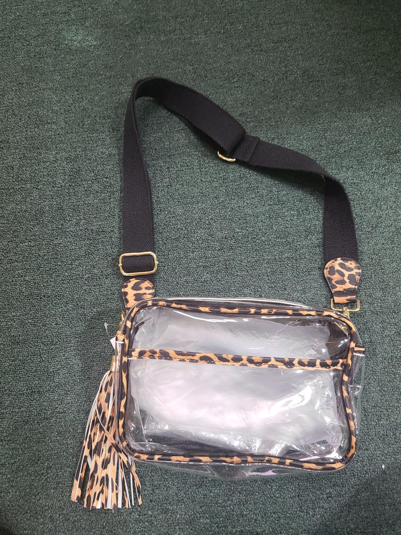 Puede incluir: Bolso cruzado transparente con correa negra y detalles de estampado de leopardo. El bolso tiene forma rectangular con cierre de cremallera y un adorno de borla. Se ve herraje de color dorado.