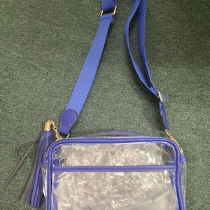 Puede incluir: Un bolso cruzado transparente con una correa y ribete azules ajustables. El bolso tiene forma rectangular con cierre de cremallera y una borla azul. Los herrajes dorados acent&uacute;an la correa y la cremallera.