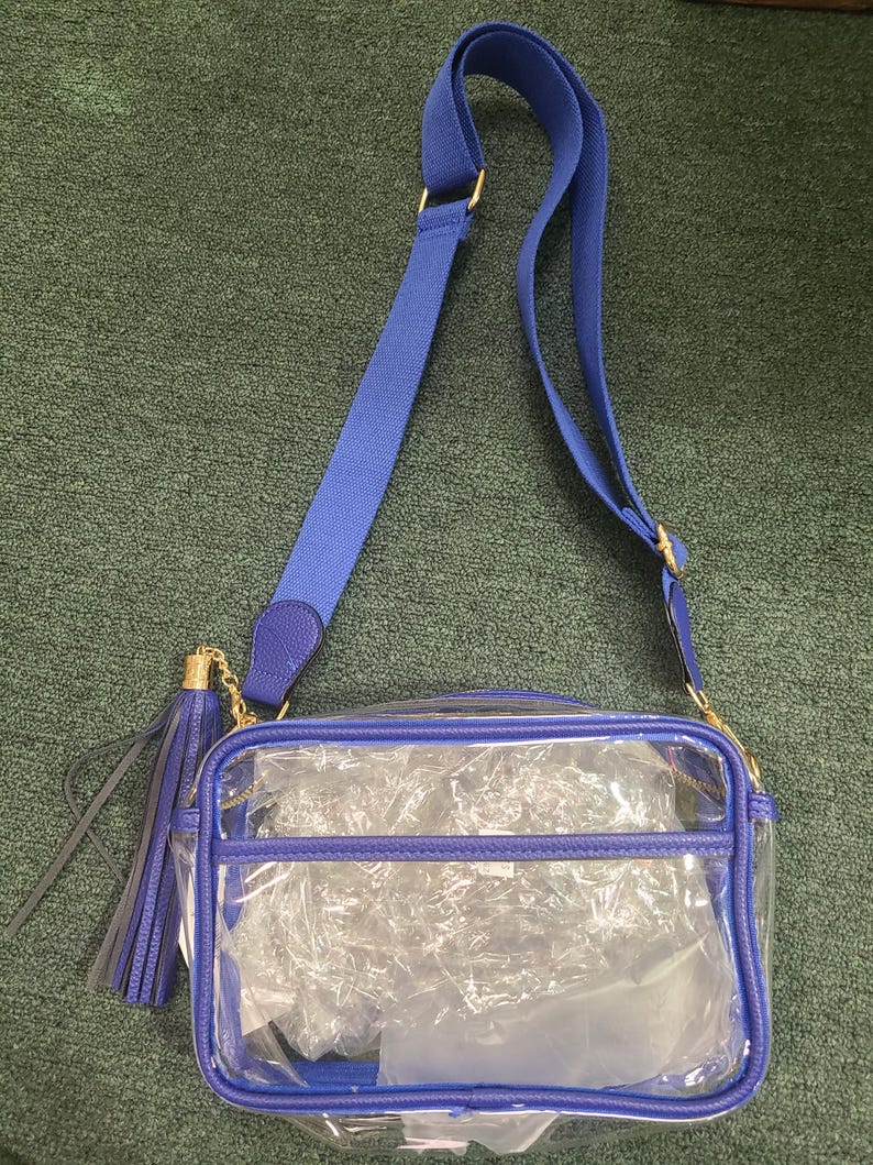 Puede incluir: Un bolso cruzado transparente con una correa y ribete azules ajustables. El bolso tiene forma rectangular con cierre de cremallera y una borla azul. Los herrajes dorados acent&uacute;an la correa y la cremallera.