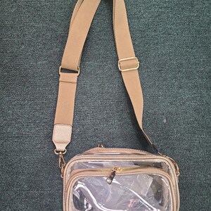 May include: A clear crossbody bag with a tan adjustable strap and gold-colored hardware. The bag has a rectangular shape with a zippered main compartment and a smaller front compartment. The bag is made of clear plastic and tan faux leather.