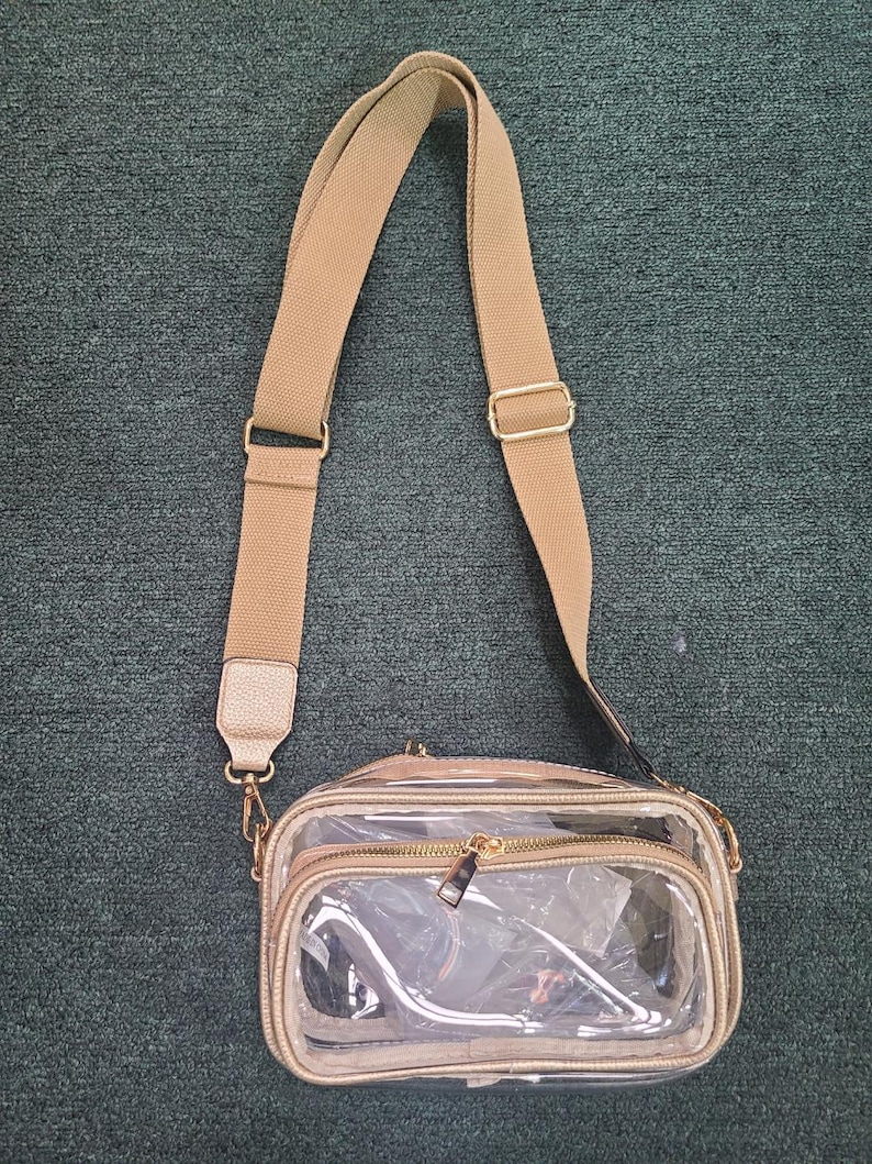 May include: A clear crossbody bag with a tan adjustable strap and gold-colored hardware. The bag has a rectangular shape with a zippered main compartment and a smaller front compartment. The bag is made of clear plastic and tan faux leather.