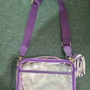 Puede incluir: Un bolso cruzado rectangular transparente con ribetes morados y una correa ajustable a juego. El bolso tiene una cremallera y herrajes dorados, y una borla blanca y morada. El bolso est&aacute; dise&ntilde;ado para ser transparente.