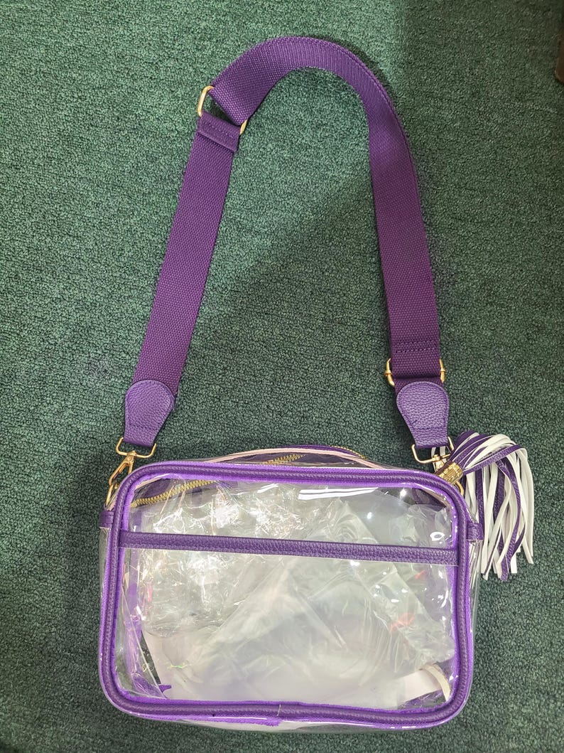 Puede incluir: Un bolso cruzado rectangular transparente con ribetes morados y una correa ajustable a juego. El bolso tiene una cremallera y herrajes dorados, y una borla blanca y morada. El bolso est&aacute; dise&ntilde;ado para ser transparente.