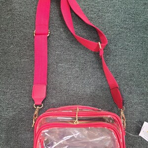 May include: A red crossbody bag with a clear front panel and a matching red adjustable strap. The bag has gold-colored hardware and a zippered closure. The strap is approximately 40 inches long.