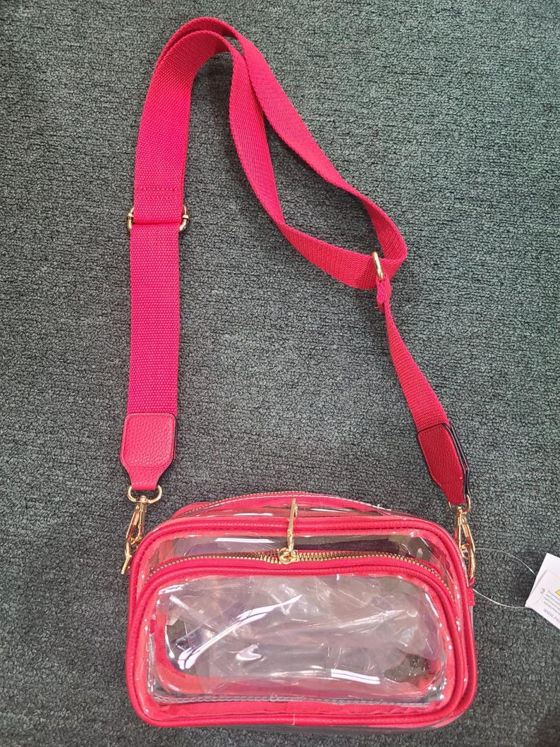 May include: A red crossbody bag with a clear front panel and a matching red adjustable strap. The bag has gold-colored hardware and a zippered closure. The strap is approximately 40 inches long.