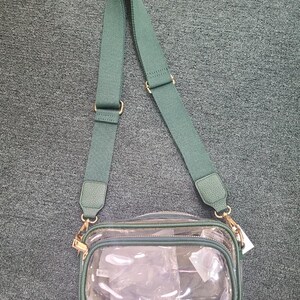 May include: Clear crossbody bag with a green adjustable strap and trim. The bag has a rectangular shape with a zippered top and gold-colored hardware. The bag is empty and ready to be filled.