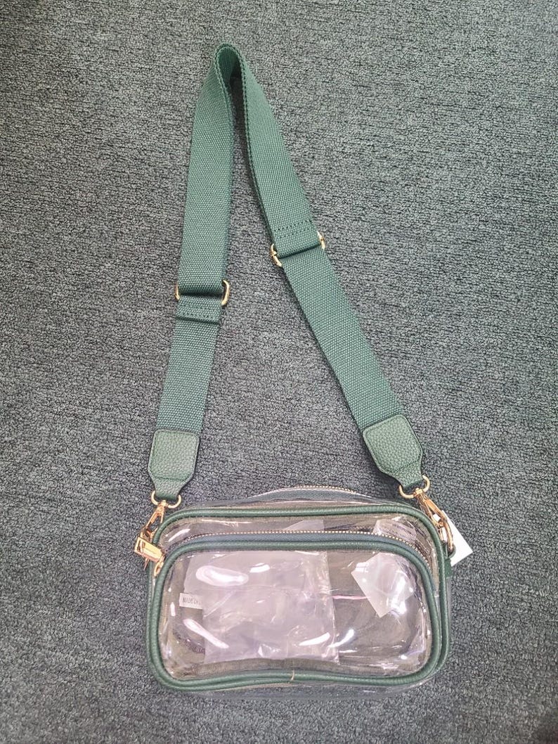 May include: Clear crossbody bag with a green adjustable strap and trim. The bag has a rectangular shape with a zippered top and gold-colored hardware. The bag is empty and ready to be filled.