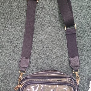 May include: A clear crossbody bag with a black adjustable strap and gold-colored hardware. The bag has a rectangular shape with a zippered closure and a small, clear front pocket. The strap is made of a durable, woven material.