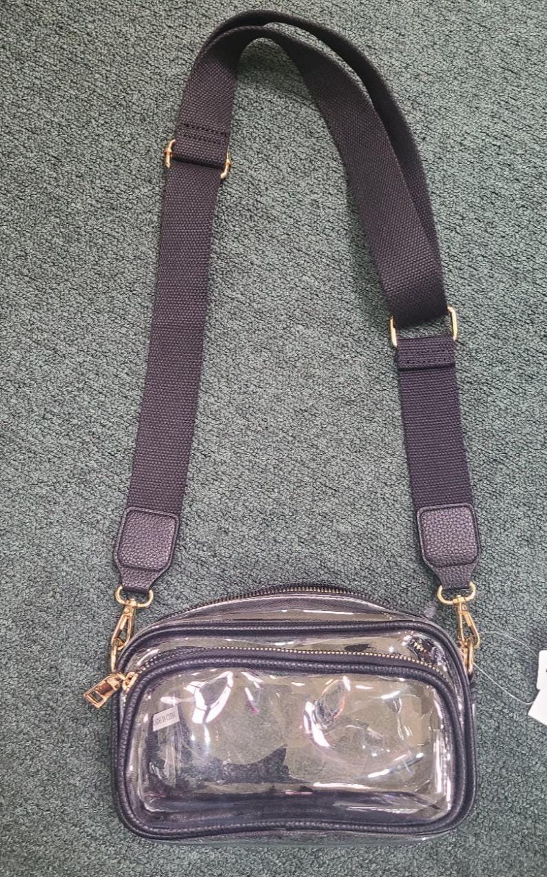 May include: A clear crossbody bag with a black adjustable strap and gold-colored hardware. The bag has a rectangular shape with a zippered closure and a small, clear front pocket. The strap is made of a durable, woven material.