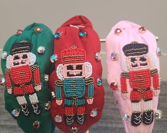 Op de afbeelding: Drie kerstthema-hoofdbanden met kralen-nootkraker-ontwerpen. De hoofdbanden zijn groen, rood en roze. Elke hoofdband heeft een nootkraker in een andere kleur met strass.