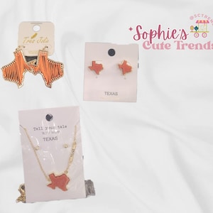 May include: Three sets of Texas-themed jewelry: a pair of orange and gold earrings shaped like the state of Texas, a pair of gold stud earrings with a small orange Texas outline, and a gold necklace with a pendant shaped like the state of Texas and a small charm that says "Tell your tale" with a smaller charm that says "Texas".