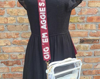 May include: Clear crossbody bag with a white zip and a maroon and white beaded strap that says "GIG 'EM AGGIES". The bag has a white tassel and a gold chain. The bag is worn over a black dress.