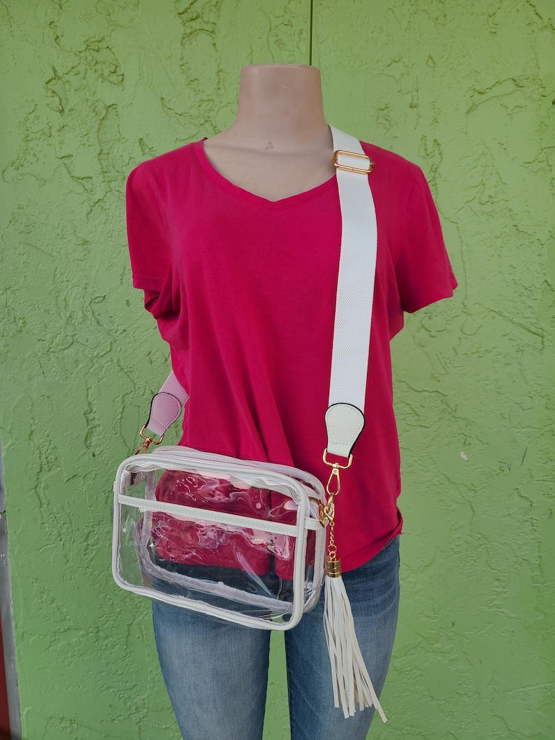 Puede incluir: Un bolso cruzado transparente con una correa blanca y herrajes dorados. El bolso tiene un ribete blanco y una borla blanca. El bolso se lleva sobre una camiseta rosa con cuello en V y vaqueros azules.