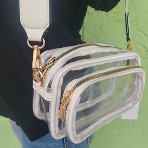 May include: Clear crossbody bag with a white and black patterned design. The bag has a white adjustable strap and gold-colored zippers and hardware. The bag has two compartments and a small tag attached.