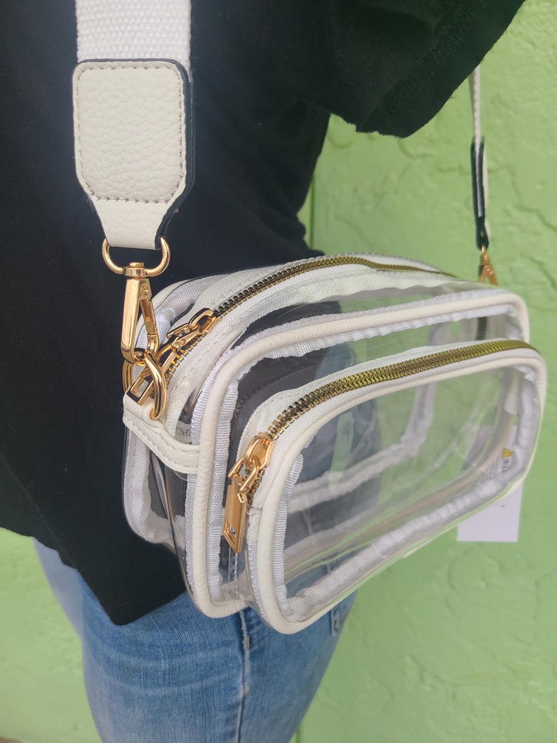 May include: Clear crossbody bag with a white and black patterned design. The bag has a white adjustable strap and gold-colored zippers and hardware. The bag has two compartments and a small tag attached.