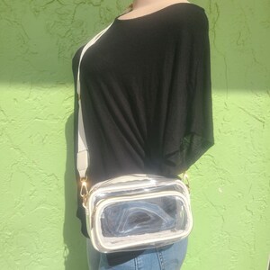 May include: A clear, rectangular crossbody bag with a white trim and a white adjustable strap. The bag has a gold zipper and hardware. The mannequin is wearing a black top and blue jeans.