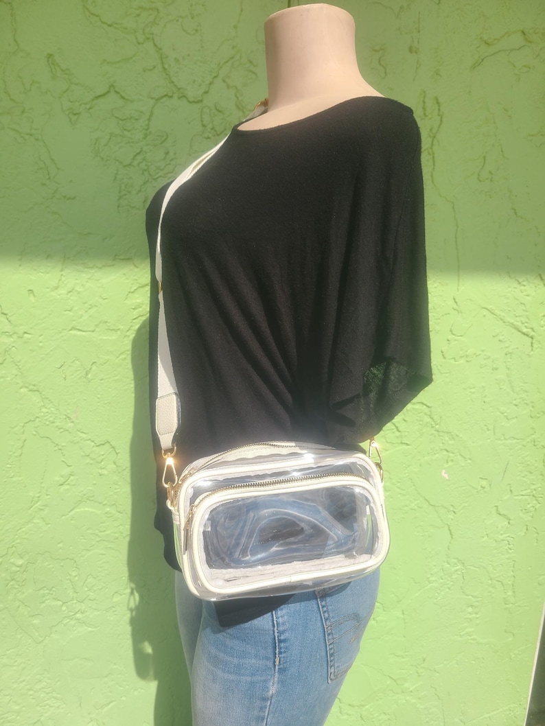 May include: A clear, rectangular crossbody bag with a white trim and a white adjustable strap. The bag has a gold zipper and hardware. The mannequin is wearing a black top and blue jeans.
