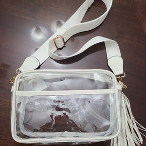Puede incluir: Un bolso cruzado transparente con correa y ribete blancos. El bolso tiene forma rectangular con una borla blanca y herrajes dorados. La correa ajustable es blanca.