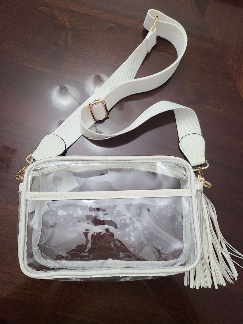 Puede incluir: Un bolso cruzado transparente con correa y ribete blancos. El bolso tiene forma rectangular con una borla blanca y herrajes dorados. La correa ajustable es blanca.