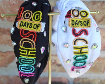 Könnte beinhalten: Zwei Stirnbänder, eines in Schwarz und eines in Weiß, werden auf einem goldenen Ständer präsentiert. Beide Stirnbänder zeigen den Text "100 DAYS OF SCHOOL" in farbigen Perlenbuchstaben und sind mit schillernden Strasssteinen verziert. Das schwarze Stirnband hat einen Regenbogenverlauf, das weiße ein rot-gelbes Farbschema.