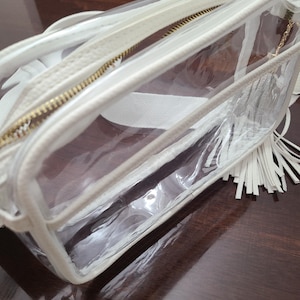 Puede incluir: Un neceser rectangular transparente con ribete blanco y cremallera dorada. La bolsa tiene una borla blanca y una correa blanca. La bolsa est&aacute; hecha de pl&aacute;stico transparente y est&aacute; parcialmente abierta.