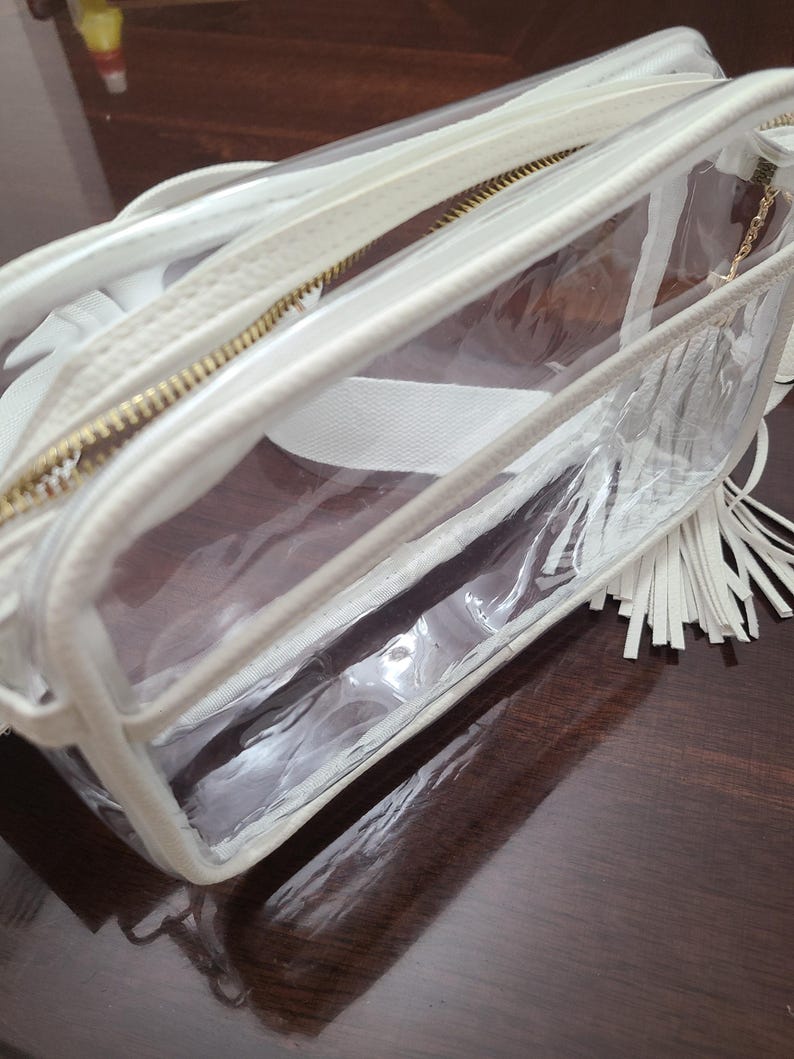 Puede incluir: Un neceser rectangular transparente con ribete blanco y cremallera dorada. La bolsa tiene una borla blanca y una correa blanca. La bolsa est&aacute; hecha de pl&aacute;stico transparente y est&aacute; parcialmente abierta.