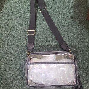 Puede incluir: Un bolso cruzado transparente con una correa negra ajustable y herrajes dorados. El bolso tiene un ribete negro y una borla. El interior del bolso est&aacute; lleno de material de embalaje blanco.