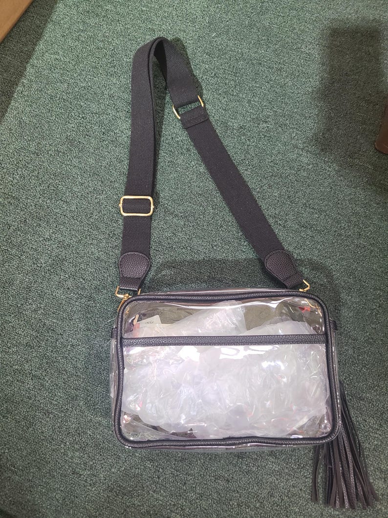Puede incluir: Un bolso cruzado transparente con una correa negra ajustable y herrajes dorados. El bolso tiene un ribete negro y una borla. El interior del bolso est&aacute; lleno de material de embalaje blanco.