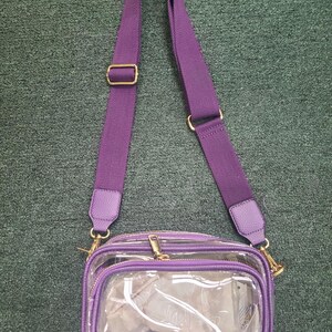 May include: A clear crossbody bag with a purple adjustable strap and trim. The bag has a rectangular shape with a zippered main compartment and a smaller front pocket. Gold-colored hardware accents the bag.