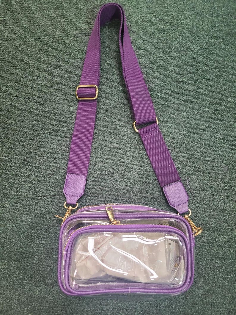 May include: A clear crossbody bag with a purple adjustable strap and trim. The bag has a rectangular shape with a zippered main compartment and a smaller front pocket. Gold-colored hardware accents the bag.