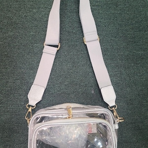 May include: A clear crossbody bag with a silver trim and a gray adjustable strap. The bag has a gold zipper and hardware. The interior of the bag is visible through the clear material.