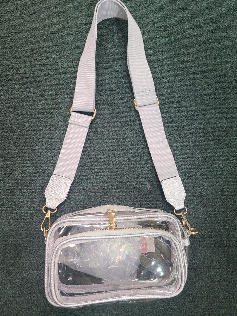 May include: A clear crossbody bag with a silver trim and a gray adjustable strap. The bag has a gold zipper and hardware. The interior of the bag is visible through the clear material.