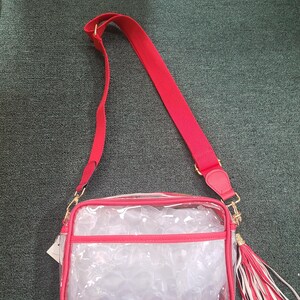 Puede incluir: Un bolso cruzado transparente con ribetes rojos y una correa para el hombro roja. El bolso tiene una cremallera y herrajes dorados, y una borla con detalles rojos y blancos. El bolso es rectangular y transparente.