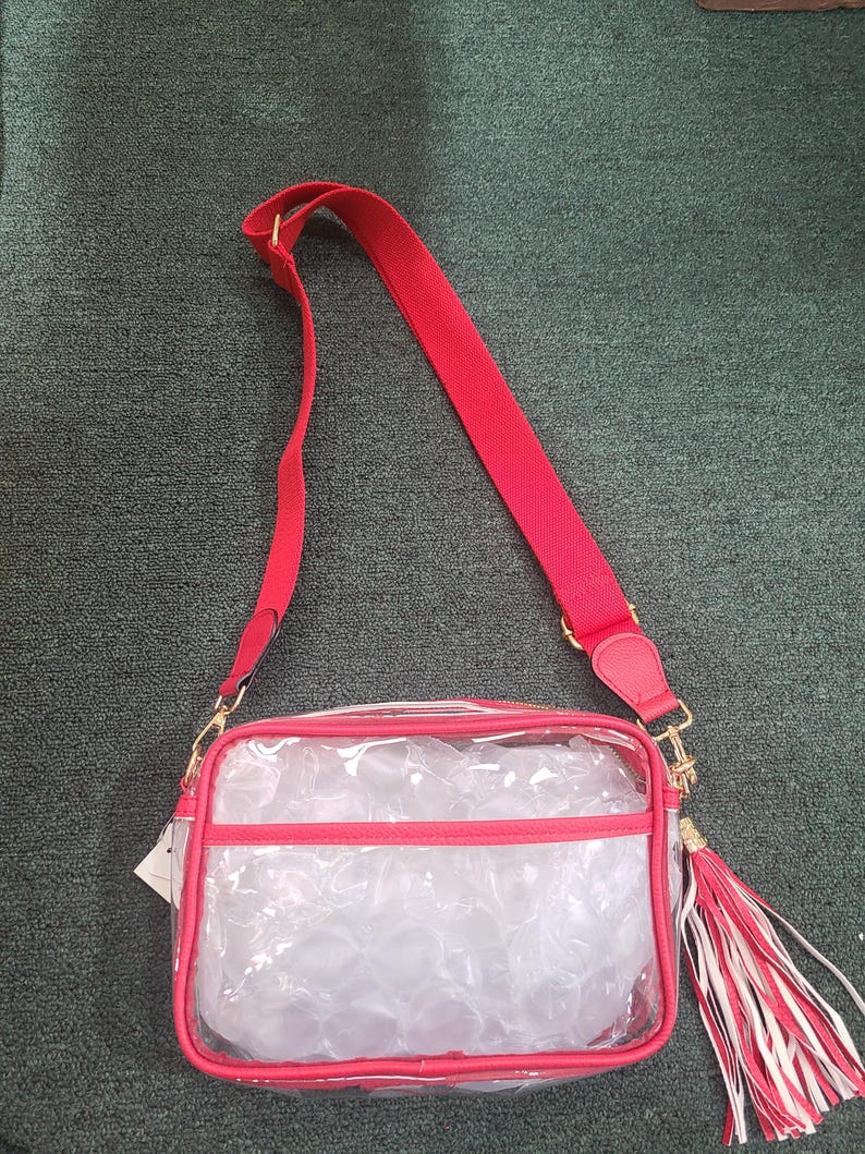 Puede incluir: Un bolso cruzado transparente con ribetes rojos y una correa para el hombro roja. El bolso tiene una cremallera y herrajes dorados, y una borla con detalles rojos y blancos. El bolso es rectangular y transparente.