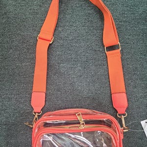 May include: A clear crossbody bag with a bright orange adjustable strap and trim. The bag has a rectangular shape with a zippered main compartment and a smaller front pocket. Gold-tone hardware accents the bag.