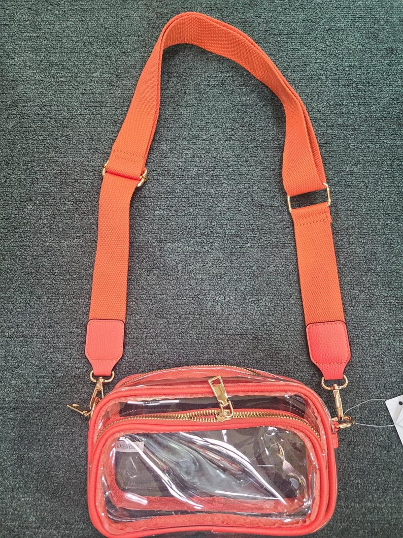 May include: A clear crossbody bag with a bright orange adjustable strap and trim. The bag has a rectangular shape with a zippered main compartment and a smaller front pocket. Gold-tone hardware accents the bag.