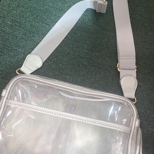 Puede incluir: Un bolso cruzado transparente y rectangular con ribete plateado y una correa ajustable gris claro. El bolso tiene cierre de cremallera y herrajes dorados. La correa es de material tejido.