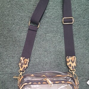 May include: Clear crossbody bag with a leopard print trim and strap accents. The bag has a black adjustable strap with gold-tone hardware. The main compartment is transparent, and the bag has a zipper closure.