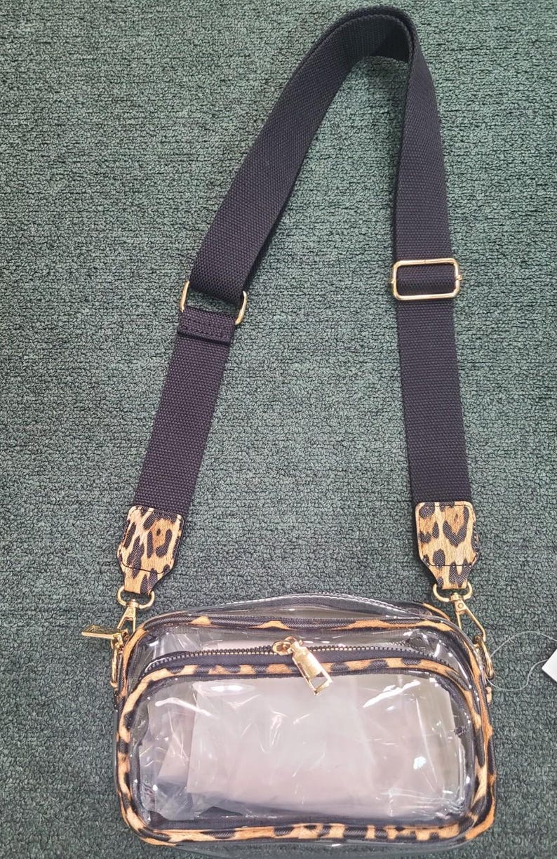 May include: Clear crossbody bag with a leopard print trim and strap accents. The bag has a black adjustable strap with gold-tone hardware. The main compartment is transparent, and the bag has a zipper closure.