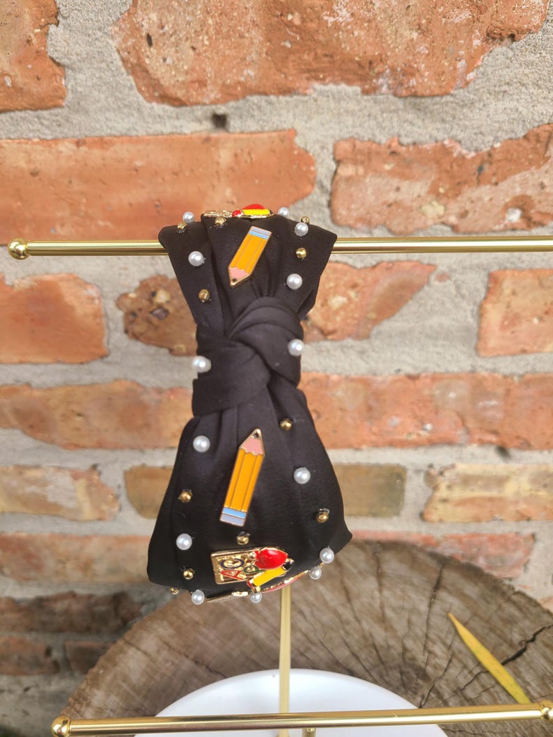 Peut inclure: Un serre-t&ecirc;te en tissu noir avec un n&oelig;ud nou&eacute; et des accents de perles. Le serre-t&ecirc;te est d&eacute;cor&eacute; d'&eacute;pingles en &eacute;mail color&eacute; repr&eacute;sentant des fournitures scolaires, notamment des crayons, une r&egrave;gle et une pomme.