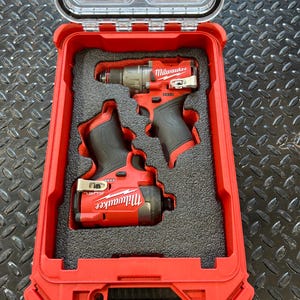 May include: Two red Milwaukee power tools, a drill and a driver, are stored in a red hard case with a clear lid. The tools are nestled in black foam cutouts.