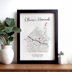 Op de afbeelding: Een zwart ingelijst afdruk met een witte achtergrond. De afdruk toont een kaart van een stad in de vorm van een huis. De tekst "Oliver & Hannah" staat bovenaan de afdruk. Er zijn rode harten verspreid over de kaart. De tekst "261 Roman Road" en "28 October 2024" staat onderaan de afdruk.