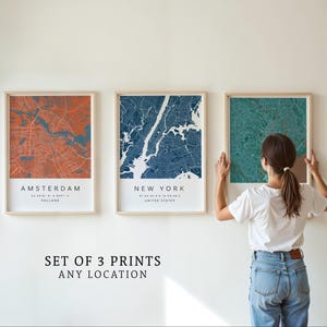 Peut inclure: Ensemble de trois impressions encadrées de cartes de villes en orange, bleu et vert d'eau. La carte d'Amsterdam est orange, celle de New York est bleue et la troisième est vert d'eau. Les impressions sont sur un mur blanc. Texte : "SET OF 3 PRINTS ANY LOCATION."