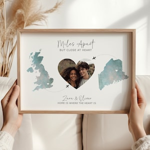 May include: A framed print with a watercolour illustration of two maps connected by a heart shape. The heart contains a photo of a couple. The text reads "Miles Apart But Close At Heart" and "Zara & Oliver Home Is Where The Heart Is."