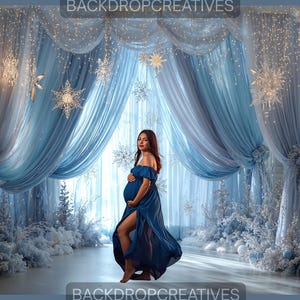 May include: A pregnant person in a flowing, off-the-shoulder blue dress poses in front of a winter-themed backdrop. The backdrop features light blue curtains, snowflake decorations, and small, white Christmas trees. The text "BACKDROPCREATIVES" is visible.