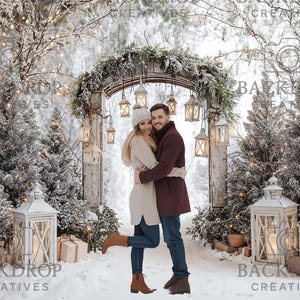 May include: A winter-themed backdrop featuring a couple embracing in a snowy scene. The backdrop includes a wooden arch adorned with greenery and hanging lanterns. White lanterns and snow-covered trees frame the couple, creating a festive atmosphere.