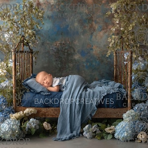 May include: A newborn baby sleeps peacefully on a small wooden bed, draped with a blue blanket and pillow. The bed is surrounded by blue and white flowers and greenery, set against a textured blue and brown backdrop.