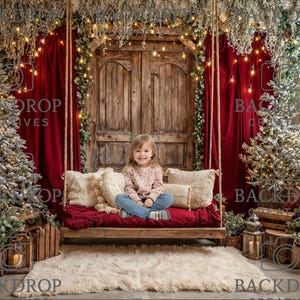 May include: A young girl sits on a wooden swing adorned with pillows and a red blanket, set against a Christmas backdrop. The scene features two Christmas trees, red curtains, and fairy lights.